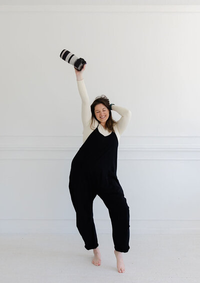 Tia Quirk, wedding photographer based in MN, holding her camera above her head while wearing a black jumpsuit in front of a white wall.