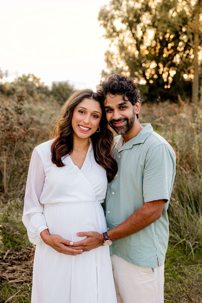 pregnant mom wearing a white dress standing in a field of tall grass during golden hour