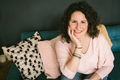 Kate sits on a teal couch, resting her head on her hand, and smiling warmly at the camera.
