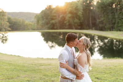 Bride and groom share a kiss with a pond and the sun setting behind them