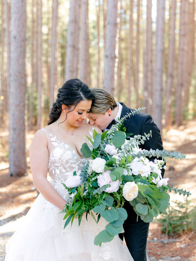 LGBTQ couple in suit and wedding gown holding a large bouquet and kissing her on the shoulder with trees in the background