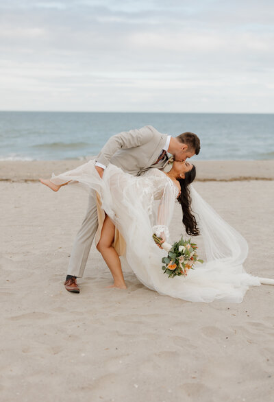A New England wedding photographer captures a groom in a light gray suit playfully dipping the bride, who wears a flowing white dress, on a sandy beach. She holds a bouquet of flowers as the ocean glistens under the partly cloudy sky—a perfect moment for elopement photography.