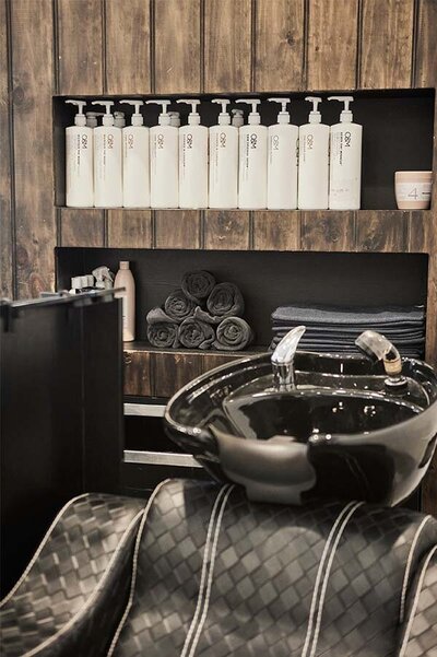 Black wash basin with a woven salon chair, surrounded by neatly arranged O&M hair products and rolled towels on wooden shelving.