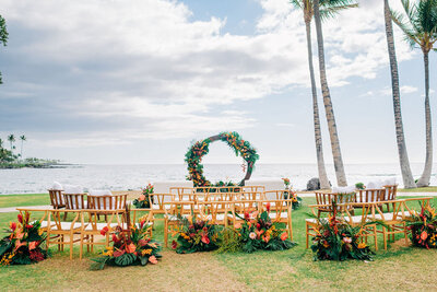 Elegant wedding ceremony setup at the Fairmont Orchid, featuring a wooden circular arch adorned with fresh tropical flowers by Grace Flowers Hawaii. A thoughtfully curated mix of chairs and loveseats creates an intimate and stylish seating arrangement for a luxury destination wedding