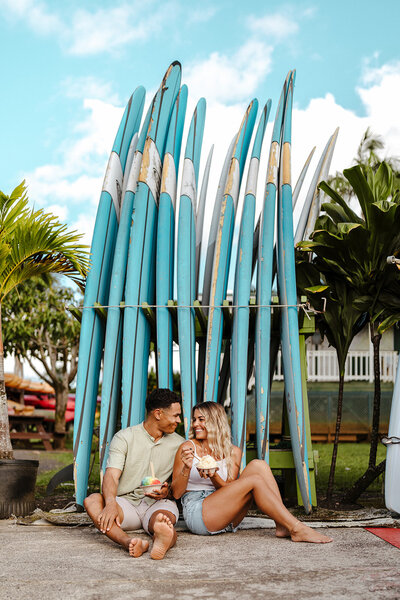 Couple sitting barefoot by a stack of surfboards in Kauai, laughing and sharing Hawaiian shave ice during their engagement session.