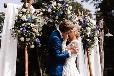 Bruiloft foto van ceremonie bruidspaar bruid en bruidegom  geeft kus zoen onder bloemenboog blauw pak kanten jurk