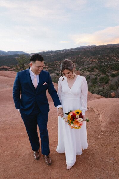 Groom and bride look down as they walk hand in hand towards the camera and the sun sets behind them.