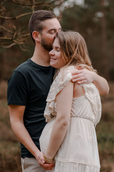 Man geeft zijn zwangere vrouw een kus op haar slaap terwijl ze dicht tegen elkaar aan staan.