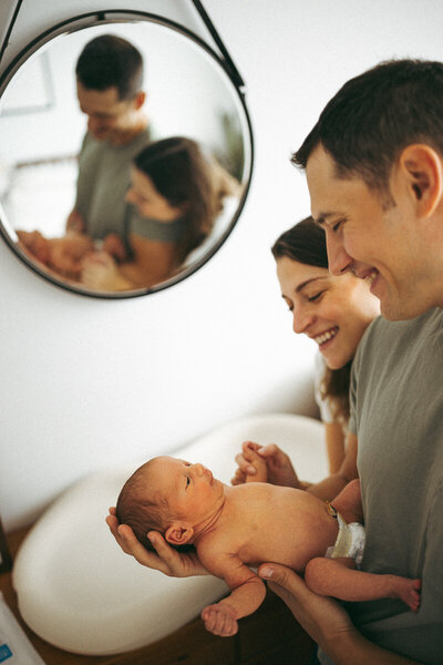 infant being held by mom and dad in the baby's room near changing table while they smile at baby in the mirror