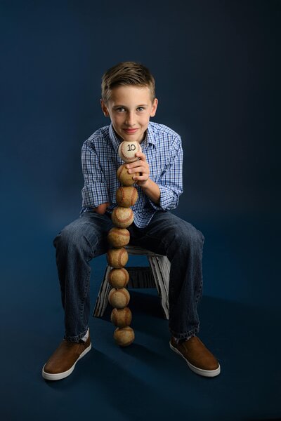 A young boy sits on a stool against a dark background, holding a tall stack of baseballs.