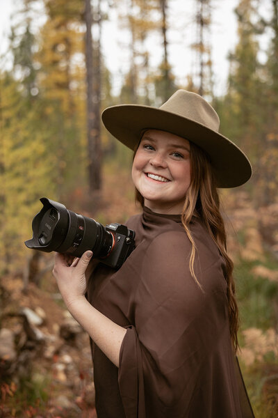 Sydney Breann, a Montana-based elopement photographer, smiles while holding her camera in a forest surrounded by autumn trees.