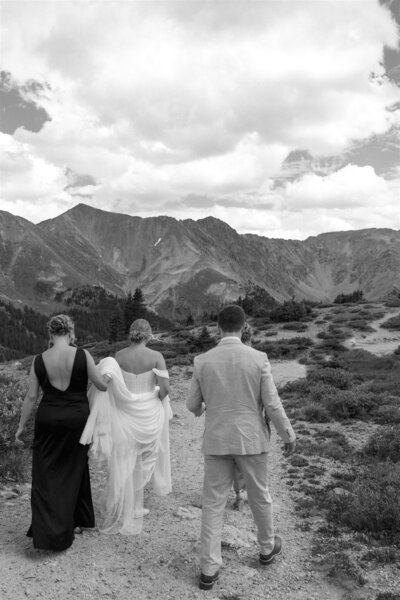 A bride, groom, and their family walking towards their ceremony spot in Colorado as the brides mom holds her dress up.