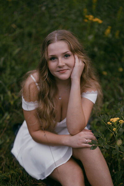 Gorgeous senior photography taken by Morgan Ashley Lynn Photography in Lake Country, WI of a girl sitting in the grass in a white dress, looking up and smiling softly with her hand on her cheek