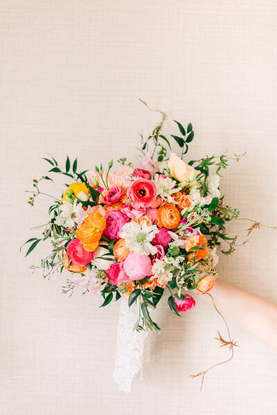 Pink, orange, white and yellow flowers with green leaves arranged whimsically set in a white container against a neutral backdrop