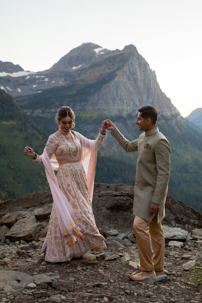 A bride and groom wearing traditional Indian wedding attire dance in the mountains of Glacier National Park.