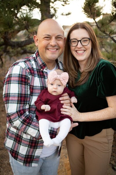Parents dressed in Christmas clothing hold their baby and look smiling at the camera.