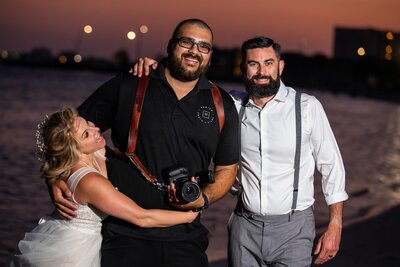 bride and groom standing with photographer while they all smile