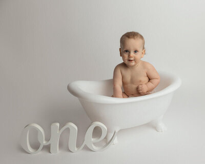 Baby Atlas sitting in a small white clawfoot tub with cake frosting on their face after a cake smash session.