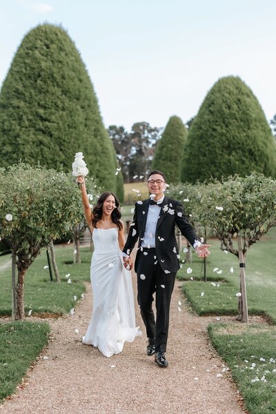 A bride and groom walking in the garden with white rose petals in the air.