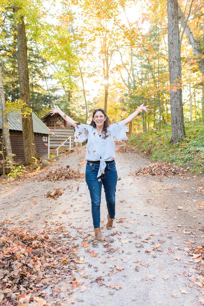 A woman standing on a path with cabins in the background. She is throwing leaves in the air and smiling.