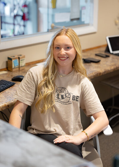 Chiropractic assistant smiling in clinic setting wearing a tan Busy Bee Chiropractic shirt.