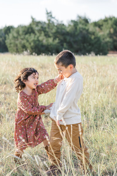 A family of four poses in a park for their family portraits in Austin, TX. They look and smile at the camera. The mom holds one of their sons in her arms while his brother stands in front of his father.
