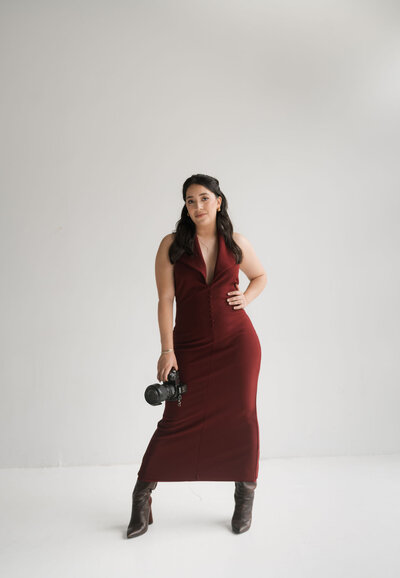 Photographer in a fitted maroon dress holds a camera, stands confidently against a plain white background. She wears brown boots, exuding a poised and professional aura.