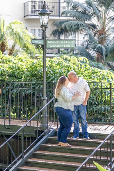 Husband and wife photography team shares a tender kiss on a staircase adorned with vibrant plants and a sign indicating nearby amenities, reflects the romantic essence of North Carolina wedding photography.