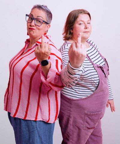 Two people stand back-to-back in a studio, smirking as they hold up their middle fingers toward the camera. Pippa on the left wears glasses, a red-and-pink striped blouse, and jeans; Lucy on the right wears a black-and-white striped top with dusty-purple dungarees.