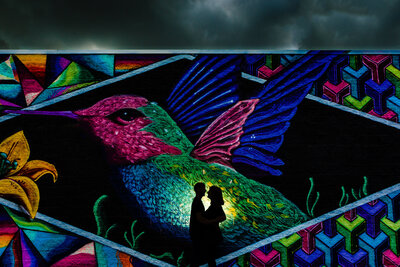 A silhouette of an engaged couple in front of a large mural of a colorful bird in Toledo Ohio