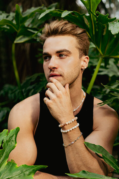young man posing in a portrait with large green plants placed behind him wearing white pearl jewelry