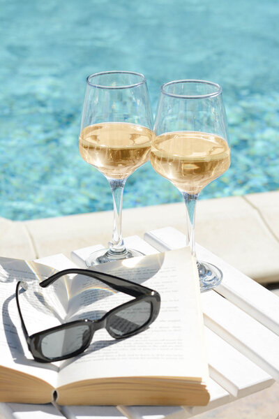 Two glasses of white wine resting on an open book beside a pool, with sunglasses placed on top of book.