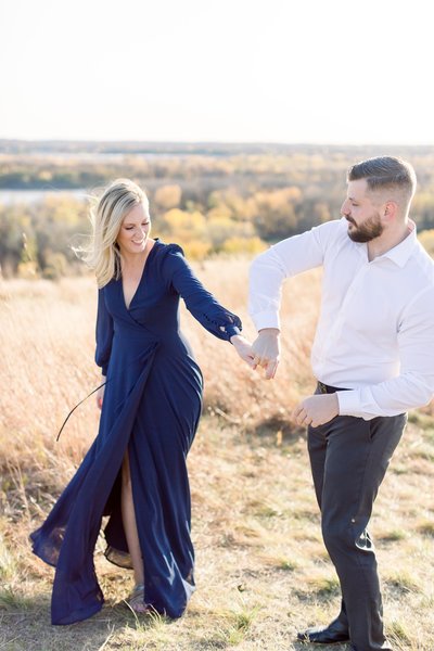 Man and woman holds hands while walking through field for engagement photography sesssion