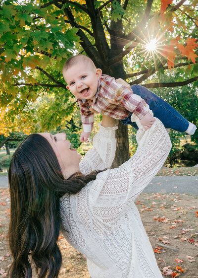 Lifestyle photo of mother holding her laughing son in the air at an Essex County, New Jersey park in the fall.