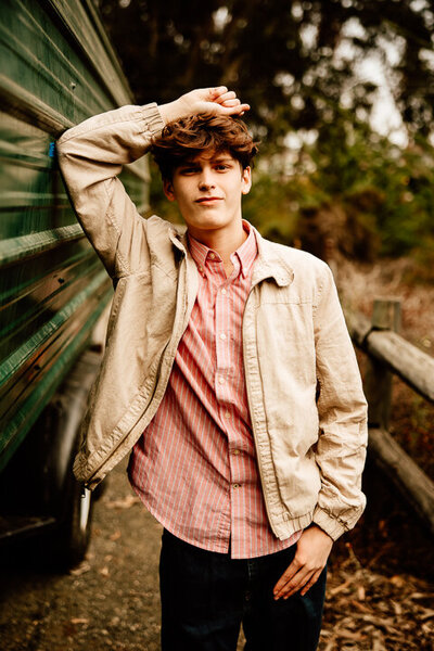 High school senior boy leaning up against a deep green trailer.  He is wearing a red shirt and tan Jacket and has one hand laying across the top of his head with his other hand in his pocket.  He is looking at the camera with a slight smirk.