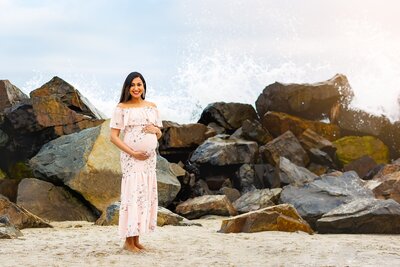 Woman in light pink gown posing for maternity portrait and looking at camera with waves splashing against rocks on the beach in the backdrop