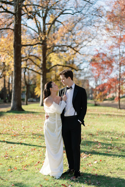 Bride in an elegant off-shoulder gown and groom in a black tuxedo standing in a sunlit park with vibrant autumn foliage.