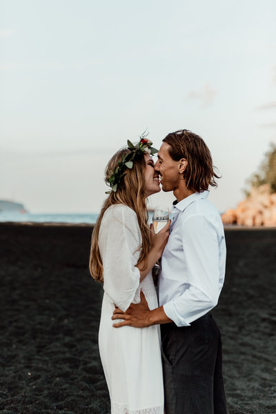 bride and groom holding champagne smiling going in for a kiss on a black sand beach
