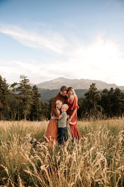 Family of four hugging each other in tall Colorado summer grass with Pikes Peak in the background