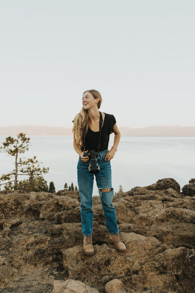 California elopement photographer Kasey Mantiply wearing blue jeans black top and hiking boots with camera around neck looking off to the left standing on rocks in front of Lake Tahoe