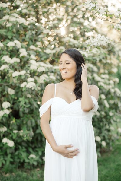 Pregnant mom holding belly in white dress in front of flowers by Northern Virginia Maternity Photographer