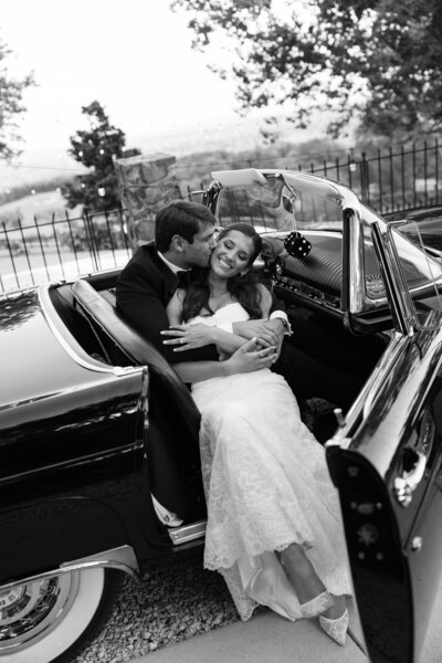 Black-and-white photo of groom kissing bride on cheek while sitting in a vintage. antique car on their wedding day.