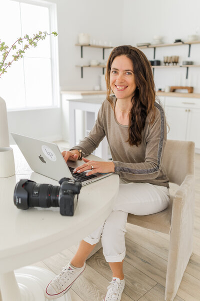 woman sitting in her family room working on her computer with her camera next to her with white and beige colors