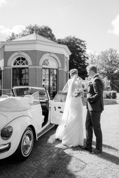 Een zwart-wit foto van een bruidspaar dat poseert naast een vintage auto voor een charmante trouwlocatie.