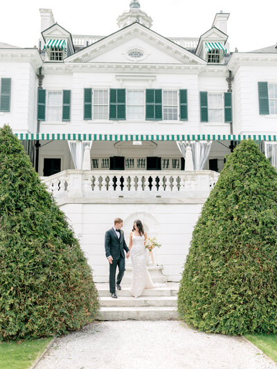 Bride and groom walk down the stairs behind The Mount Edith Wharton's Home holding hands