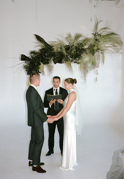 Bridal couple in suit and high neck wedding dress stand together at the alter with a green mossy backdrop during ceremony