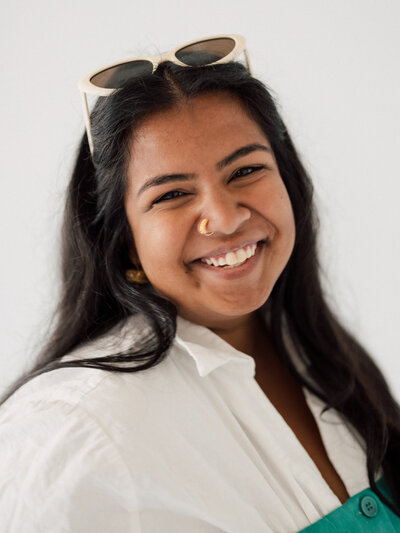 Headshot of Neha Balachandran, the owner of Jeevan Portraits Photography smiling wearing a white shirt with sunglasses on her head