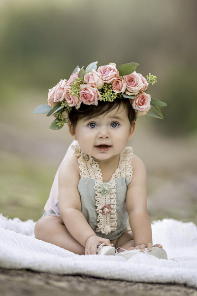 Young six month old baby sitting on a white blanket on a trail at a park outdoors while wearing a pink rose floral crown on her head and wearing a mint green romper with fabric floral embellishments added with some crochet details for trim