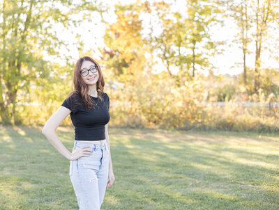 high school female senior with long dark hair, standing in grass with trees behind her, hand on her hip, turned to the side, smiling at the camera with black glasses, blue jeans, and a short sleeve black shirt