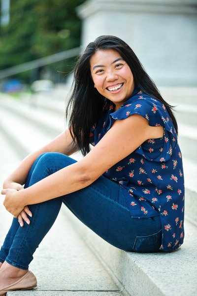 Headshot of Diana Wei Fang of The Finer Points. Diana is sitting on concrete steps with her knees up and her arms around her knees. She's smiling at the camera.